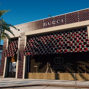 Puede incluir: Vista exterior de una tienda Gucci con el logotipo de la marca en un lugar destacado. El edificio presenta una fachada estampada con detalles en rojo y negro. Las palmeras y un cielo azul claro proporcionan un telón de fondo soleado.