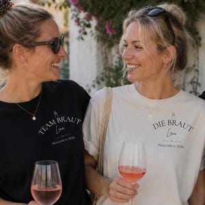 May include: Two black t-shirts and one cream t-shirt featuring the text Team Braut or Die Braut, the name Lena, and Mallorca 2026. Each shirt displays a small graphic of two champagne glasses above the text. The shirts are paired with white pants.