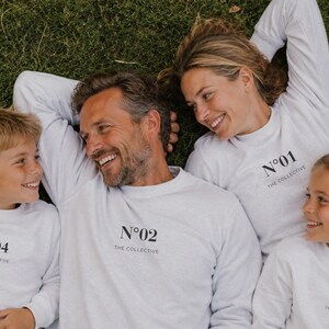 May include: Four white long-sleeve crewneck sweatshirts featuring black text on the chest. Each sweatshirt displays a unique number from N°01 to N°04 above the words THE COLLECTIVE in a clean, sans-serif font centered on the upper torso area.