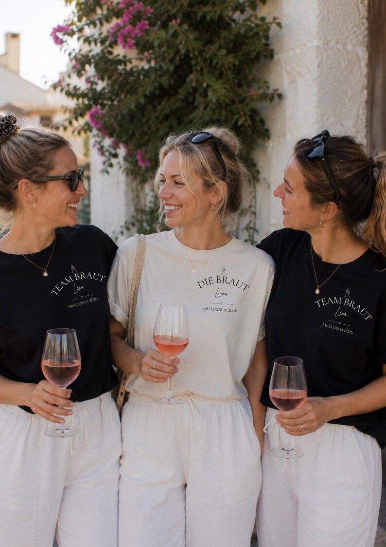 May include: Two black t-shirts and one cream t-shirt featuring the text Team Braut or Die Braut, the name Lena, and Mallorca 2026. Each shirt displays a small graphic of two champagne glasses above the text. The shirts are paired with white pants.