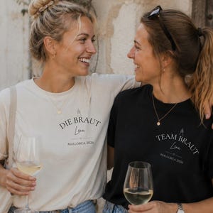 May include: A cream-colored t-shirt with the black text DIE BRAUT Lena MALLORCA 2026 and a black t-shirt with the white text TEAM BRAUT Lena MALLORCA 2026, both featuring a small graphic of a bride and a heart icon on the chest area.