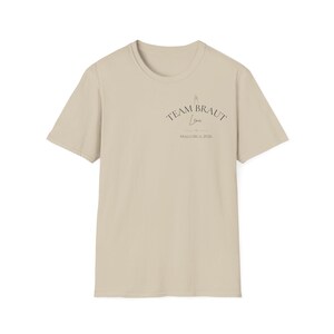 May include: A beige short-sleeve t-shirt featuring dark text on the upper left chest. The text reads TEAM BRAUT, followed by the name Lena in script, a small heart icon, and the text MALLORCA 2026. The shirt has a crew neckline and a plain back.