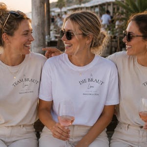 May include: Three short-sleeved crew neck t-shirts in white and beige. The two outer shirts feature the text 'TEAM BRAUT', 'Lena', and 'MALLORCA 2026'. The center shirt features the text 'DIE BRAUT', 'Lena', and 'MALLORCA 2026' in black lettering.