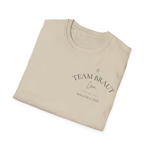 May include: A beige t-shirt featuring a dark graphic print on the chest. The text reads TEAM BRAUT, Lena, and MALLORCA 2026, accompanied by a small line drawing of two clinking champagne glasses and a tiny heart symbol centered below the name.