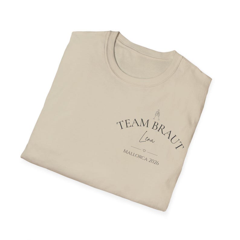 May include: A beige t-shirt featuring a dark graphic print on the chest. The text reads TEAM BRAUT, Lena, and MALLORCA 2026, accompanied by a small line drawing of two clinking champagne glasses and a tiny heart symbol centered below the name.