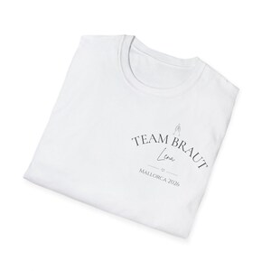 May include: A white t-shirt featuring black text that reads TEAM BRAUT, with the name Lena written in a script font below it, followed by a small heart icon and the text MALLORCA 2026. A small line drawing of two clinking glasses is above the text.