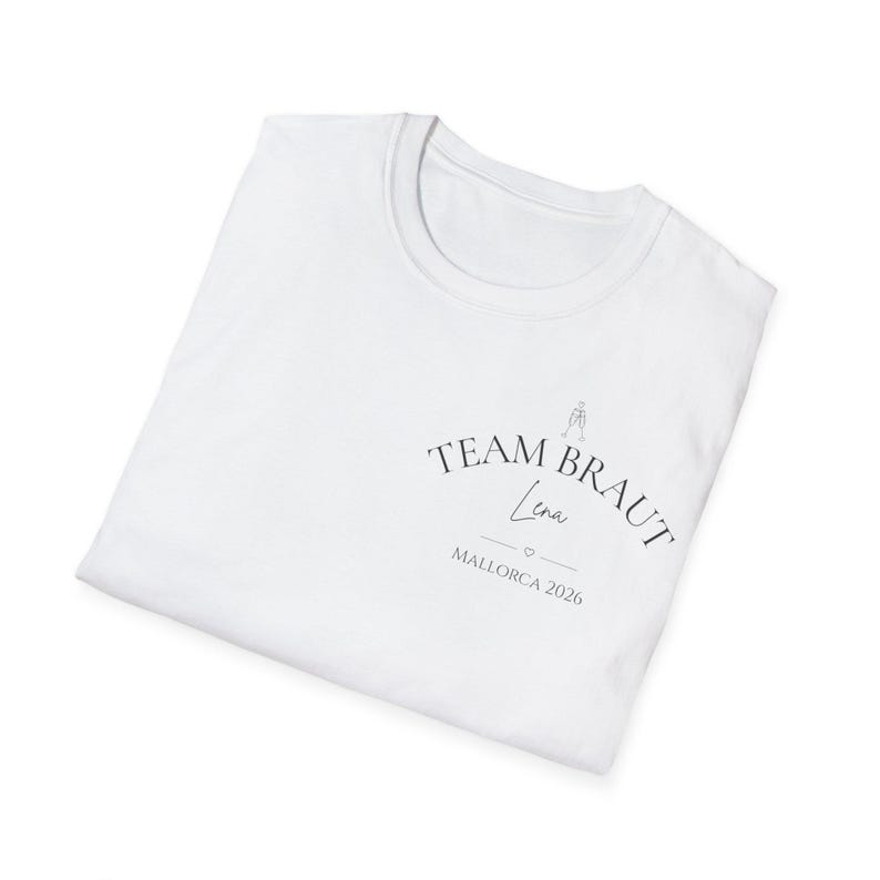 May include: A white t-shirt featuring black text that reads TEAM BRAUT, with the name Lena written in a script font below it, followed by a small heart icon and the text MALLORCA 2026. A small line drawing of two clinking glasses is above the text.
