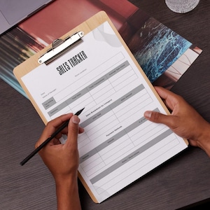 May include: A wooden clipboard with a printed "SALES TRACKER" form. The form includes sections for sales data, payment methods, and notes. A person is holding the clipboard and a black pen, ready to fill out the form.