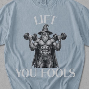 Pode incluir: Camiseta azul claro com as palavras "LIFT YOU FOOLS" em branco. O desenho mostra um mago musculoso levantando halteres. A camiseta tem gola redonda e mangas curtas. O logotipo da marca Comfort Colors é visível.