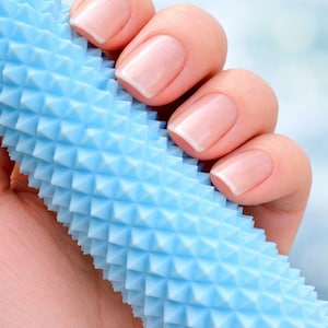 May include: A light blue, textured massage roller with a repeating pyramid pattern. The roller is held in a hand with manicured nails, showcasing a classic French manicure. The background is a soft, blurred gradient of light blue and white.
