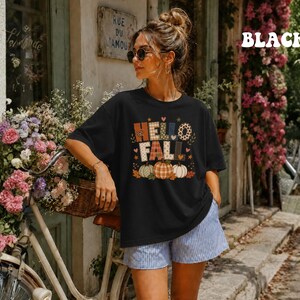 Puede incluir: Una camiseta negra de manga corta con un estampado gráfico que incluye el texto HELLO FALL en una mezcla de patrones de cuadros, leopardo y colores sólidos. El diseño presenta tres calabazas en naranja, blanco y verde, rodeadas de hojas de otoño.