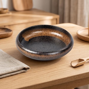 May include: A round ceramic bowl with a speckled black interior and a brown and black rim. The bowl sits on a wooden surface, suggesting a kitchen or dining setting. The bowl's design is simple and elegant.
