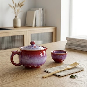 Puede incluir: Una tetera de cerámica roja y morada con una taza a juego y una cuchara de madera sobre una mesa de madera. La tetera tiene un asa y una tapa con un pequeño pomo. La taza es pequeña y redonda.