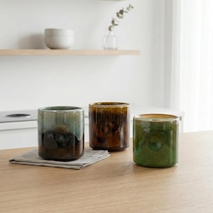 May include: Three ceramic tumblers in shades of green, brown, and black sit on a wooden surface. The tumblers have a glossy finish and a cylindrical shape. A gray linen napkin is under one of the tumblers.