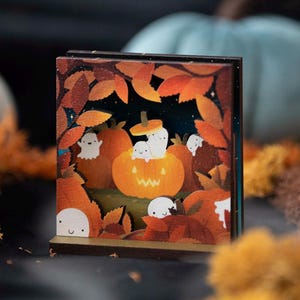 May include: A square wooden display piece featuring a colourful illustration of small white ghosts playing among orange pumpkins and autumn leaves. The artwork depicts a central jack-o'-lantern with a carved face surrounded by foliage.