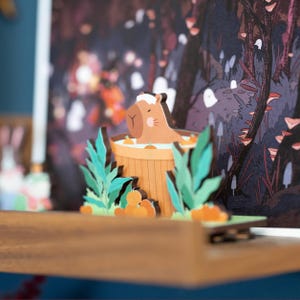 Op de afbeelding: Een grillig, gelaagd papierkunstwerk met een capibara die ontspant in een houten hot tub. De sc&egrave;ne speelt zich af in een bos met oranje accenten, groene bladeren en een vage achtergrond. De capibara is lichtbruin met een witte handdoek.
