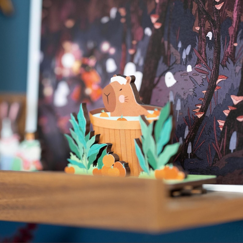 Op de afbeelding: Een grillig, gelaagd papierkunstwerk met een capibara die ontspant in een houten hot tub. De sc&egrave;ne speelt zich af in een bos met oranje accenten, groene bladeren en een vage achtergrond. De capibara is lichtbruin met een witte handdoek.