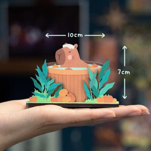 Op de afbeelding: Een grillige papieren kunstsc&egrave;ne met een capibara die ontspant in een houten hot tub. Het kunstwerk bevat groene bladeren, oranje pompoenen en meet ongeveer 10 cm breed en 7 cm hoog. De capibara draagt een witte hoed.