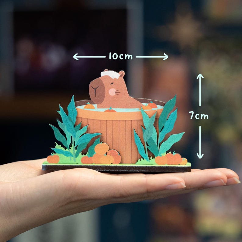 Op de afbeelding: Een grillige papieren kunstsc&egrave;ne met een capibara die ontspant in een houten hot tub. Het kunstwerk bevat groene bladeren, oranje pompoenen en meet ongeveer 10 cm breed en 7 cm hoog. De capibara draagt een witte hoed.