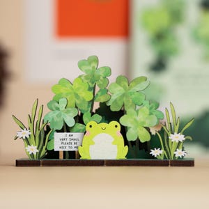 Könnte beinhalten: Ein hölzernes Frosch-Ornament mit einem Schild mit der Aufschrift "I AM VERY SMALL PLEASE BE NICE TO ME". Der Frosch ist hellgrün mit weißem Bauch, umgeben von grünen Kleeblättern, Gras und weißen Blumen. Der Sockel ist braun.