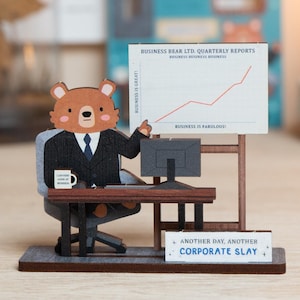 May include: A wooden desk ornament featuring a brown bear in a black suit sitting at a desk with a computer. A sign displays a rising red line graph labelled BUSINESS BEAR LTD. QUARTERLY REPORTS and BUSINESS IS FABULOUS. A small sign reads ANOTHER DAY, ANOTHER CORPORATE SLAY.