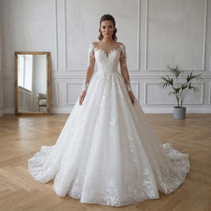 Peut inclure: Une robe de mariée blanche avec une jupe ample et des manches longues. Le corsage présente des détails en dentelle délicate et l'encolure est une illusion transparente. La robe a une traîne fluide.