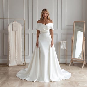 Könnte beinhalten: Ein weißes schulterfreies Brautkleid mit fließendem Rock und eng anliegendem Oberteil. Das Kleid hat einen abnehmbaren Überrock, der eine dramatische Silhouette erzeugt. Eine goldene Kleiderstange und ein Ganzkörperspiegel befinden sich im Hintergrund.