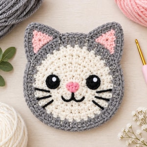 Könnte beinhalten: Ein runder, gehäkelter Untersetzer in Form eines Katzengesichts mit grauem Rand, cremefarbener Mitte und spitzen Ohren mit rosa Details. Die Katze hat gestickte schwarze Augen mit weißen Lichtpunkten, eine kleine rosa Nase und schwarze Schnurrhaare.