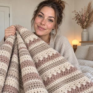 Könnte beinhalten: Eine handgehäkelte Decke mit abwechselnden horizontalen Streifen in Creme, hellem Beige und Altrosa. Das Textil zeigt ein strukturiertes Muster mit Reihen aus Stäbchen und dekorativen Büschelmaschen, die sich über das gesamte Design erstrecken.