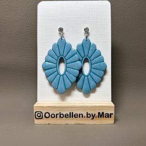 Op de afbeelding: Lichtblauwe, waaiervormige oorbellen met een ovale uitsparing in het midden. De oorbellen worden gepresenteerd op een witte kaart en een houten standaard. De tekst "Oorbellen.by.Mar" is zichtbaar.