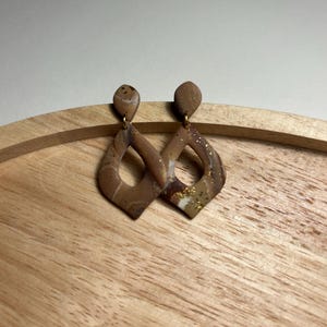 Op de afbeelding: Een paar hangende oorbellen met een ruitvormig uitgesneden ontwerp. De oorbellen zijn gemaakt van een gemarmerd bruin en beige materiaal met subtiele witte wervelingen en ingebedde goudkleurige spikkels, hangend aan kleine ronde knopjes.