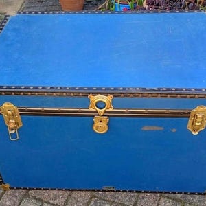 May include: A vintage blue trunk with brass hardware, including corner protectors, latches, and a central lock. The trunk has a black trim with decorative rivets. The trunk is rectangular and appears to be made of wood.