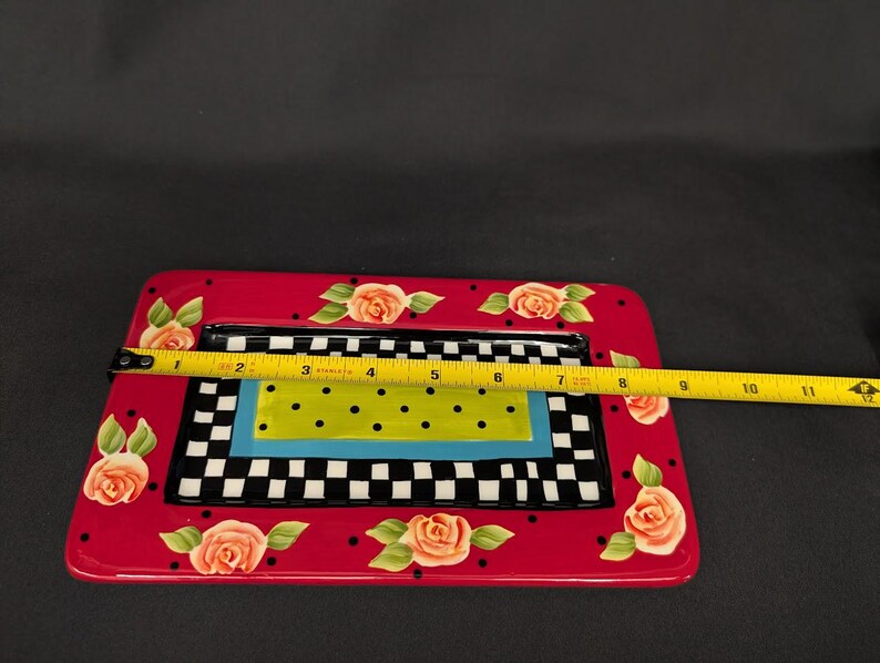 May include: A rectangular ceramic tray with a red border featuring painted orange roses and black dots. The centre has a black and white checkerboard pattern, a blue border, and a lime green rectangle with black dots. A yellow tape measure shows 28 cm.