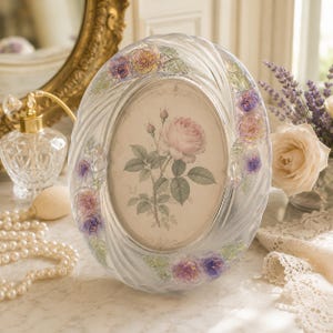 May include: An oval glass picture frame featuring a clear, swirling texture and embossed floral accents in shades of purple, pink, and green. The frame holds a central print of a single pink rose with green leaves on a cream-colored background.