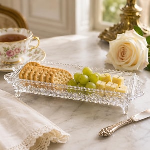 May include: A rectangular clear glass serving tray with a faceted diamond-cut pattern and four small feet. The tray holds a row of round crackers, a cluster of green grapes, and several small cubes of yellow cheese arranged on the glass surface.