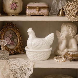 May include: A white ceramic chicken-shaped lidded dish resting in a matching basket-weave base. The chicken figure features detailed molded feathers and a comb, while the base has a textured woven pattern. The piece is a solid, opaque white.