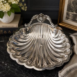 May include: A polished silver-tone serving dish shaped like a large scallop shell. The dish features a reflective metallic surface with deep, radiating fluted grooves and a decorative, ornate handle at the top edge with a shell motif detail.