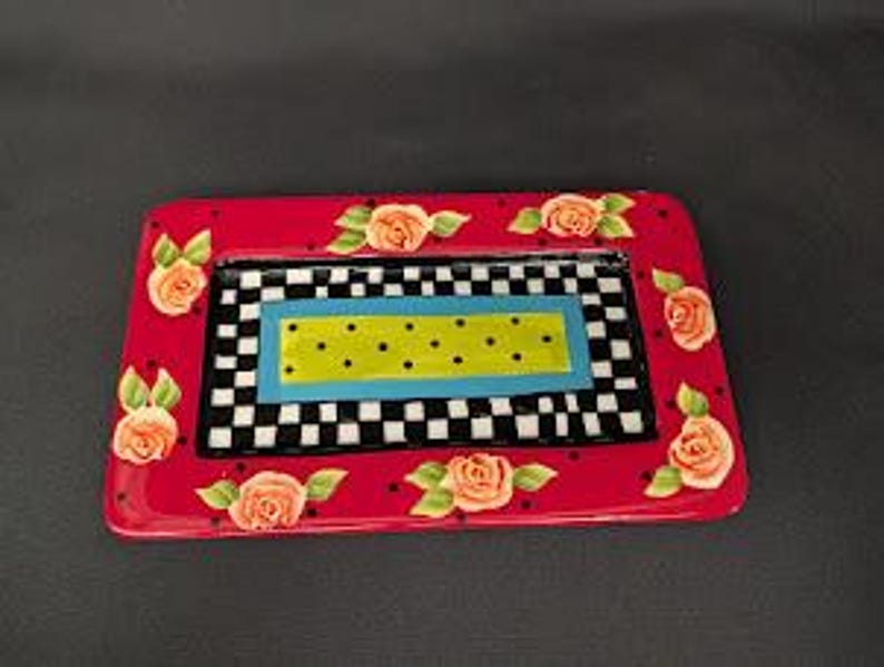 May include: A rectangular ceramic tray featuring a vibrant red border decorated with painted peach-coloured roses and green leaves. The centre displays a black and white checkered pattern surrounding a bright blue frame and a lime green dotted rectangle.