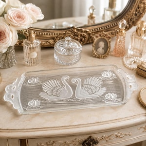 May include: A rectangular clear glass vanity tray featuring a frosted relief design of two swans facing each other, surrounded by four water lily motifs. The tray has decorative, ornate handles on both ends and a textured base surface.