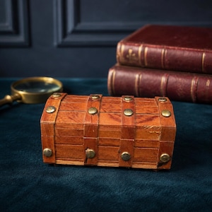 May include: A small rectangular wooden chest with a curved lid, featuring vertical wooden slats reinforced by horizontal bands and secured with round brass-colored metal studs. The chest has a natural wood grain finish and a hinged lid design.