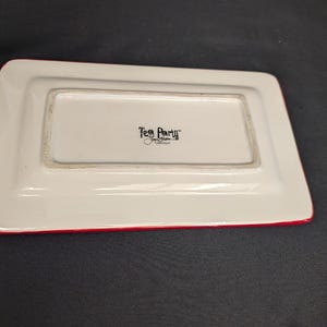 May include: A rectangular white ceramic tray with a red rim. The centre of the tray features black text that reads Tea Party by Jane Brown Collection. The tray is shown from an overhead angle, highlighting its smooth surface and clean edges.