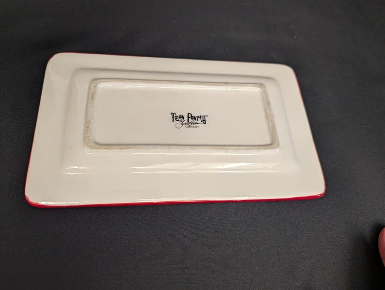 May include: A rectangular white ceramic tray with a red rim. The centre of the tray features black text that reads Tea Party by Jane Brown Collection. The tray is shown from an overhead angle, highlighting its smooth surface and clean edges.