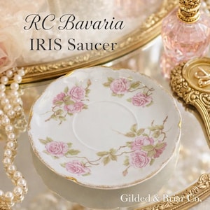May include: White RC Bavaria Iris saucer with pink rose floral design and gold trim. The saucer has a scalloped edge. Text on the saucer reads "RC Bavaria IRIS Saucer". The saucer is on a reflective surface.