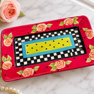 May include: A rectangular ceramic tray featuring a vibrant hot pink border decorated with painted peach-coloured roses and small black dots. The centre displays a black and white checkered pattern surrounding a bright blue and lime green rectangular panel.