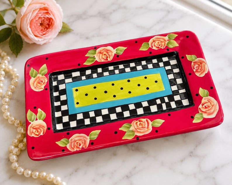 May include: A rectangular ceramic tray featuring a vibrant hot pink border decorated with painted peach-coloured roses and small black dots. The centre displays a black and white checkered pattern surrounding a bright blue and lime green rectangular panel.