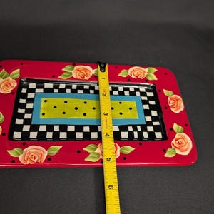 May include: A rectangular ceramic tray with a red border featuring painted peach roses and black polka dots. The centre has a black and white checkerboard pattern surrounding a blue and lime green rectangle with black dots. A yellow ruler shows a 15.24 cm width.
