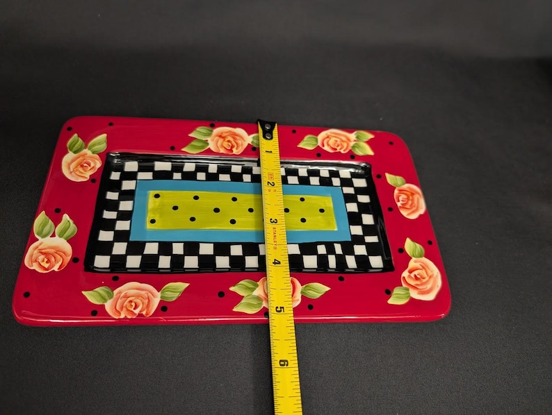 May include: A rectangular ceramic tray with a red border featuring painted peach roses and black polka dots. The centre has a black and white checkerboard pattern surrounding a blue and lime green rectangle with black dots. A yellow ruler shows a 15.24 cm width.
