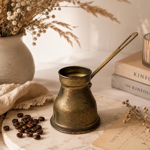 May include: A vintage brass coffee pot with a long handle sits on a marble surface. Beside it are coffee beans, a vase with dried flowers, a lit candle, and books. The pot has a classic design and a warm, aged patina.