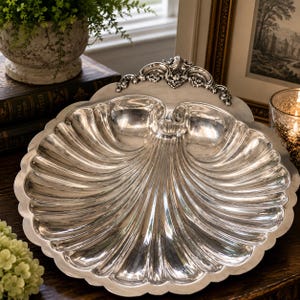 May include: A polished silver serving dish shaped like a large scallop shell. The dish features a scalloped edge and a decorative, ornate metal crest at the top center. The surface is highly reflective with deep, radiating fluted grooves.
