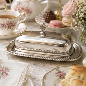 May include: A rectangular silver-plated butter dish with a removable lid featuring a decorative shell-shaped finial handle. The base and lid edges are accented with a twisted rope pattern, providing a polished, reflective surface for serving.